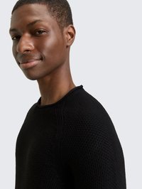 Młody mężczyzna w czarnym, teksturowanym swetrze, lekko się uśmiechający, zwrócony w lewą stronę, na tle jednolitego, jasnoszarego tła.