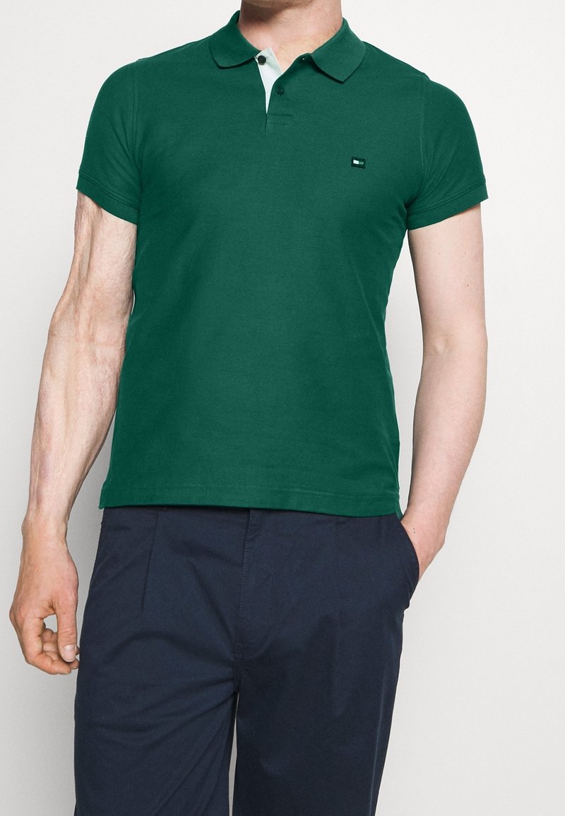 Homme portant un polo à manches courtes vert foncé avec patte de boutonnage blanche et un pantalon bleu marine, une main dans la poche, sur fond blanc.