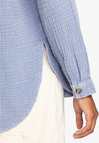 Light blue textured shirt with a wrinkled pattern, buttoned cuffs, and side slits, layered over a cream-colored garment.