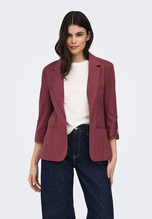 Mujer con cabello oscuro que lleva una camisa blanca, blazer burdeos con las mangas remangadas y jeans azul oscuro, de pie frente a un fondo liso.