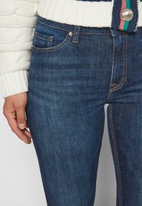 Person trägt blaue Denim-Jeans und einen weißen Strickpullover mit Knopf und grünem, pinkem sowie marineblau gestreiftem Detail am Bund.