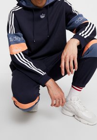 Marineblå hoodie och joggingset med vita ränder, orange och blå detaljer, Adidas-logotyp på bröstet och ärmarna, matchat med vita sneakers.