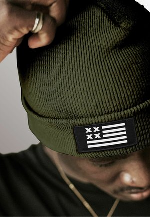 Green knitted beanie with a ribbed texture and a black patch featuring white stripes and stars, providing a contrasting detail.