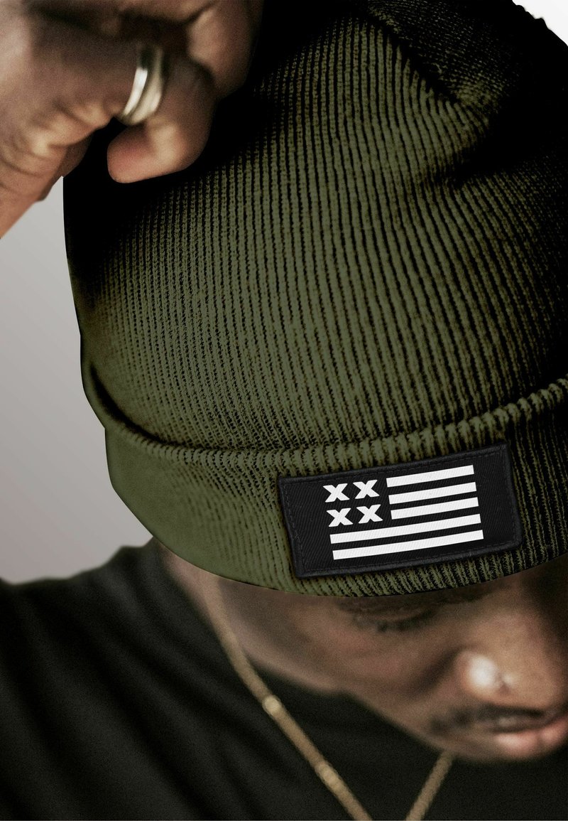 Green knitted beanie with a ribbed texture and a black patch featuring white stripes and stars, providing a contrasting detail.
