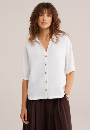 Frau mit welligen braunen Haaren, die ein weißes Hemd mit Knöpfen und Ärmeln bis zum Ellbogen sowie einen dunkelbraunen Rock trägt, steht vor einem schlichten Hintergrund.