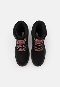 Un par de botas de invierno de ante negro con cuellos acolchados y cordones de patrón rojo y negro sobre un fondo blanco.