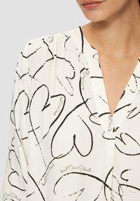 Blouse blanche avec un motif de cœurs noirs et de script, dotée d'un col en V, d'épaules plissées et d'une texture de tissu lisse.