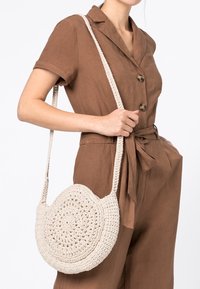 Rund, krämfärgad virkads väska med en strukturerad design och axelrem, hålls mot en brun outfit med knappar och midjeband.