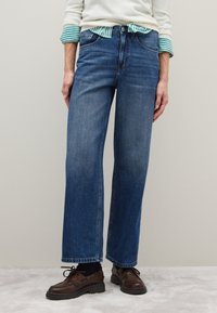 Dunkelblaue, weit geschnittene Jeans aus Denim verfügen über Fronttaschen und einen Reißverschluss, kombiniert mit braunen Schuhen und einem gestreiften Hemd darunter.