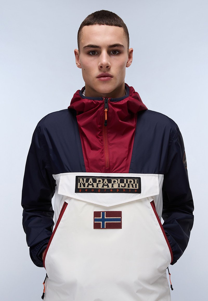 Chaqueta anorak en azul marino y blanco con acentos rojos. Presenta un bolsillo frontal, parche con el logo y una capucha. Tejido texturizado y ligero.