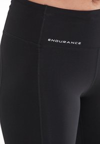 Schwarze Leggings mit hohem Bund, glatter Textur, mit weißem "ENDURANCE"-Logo auf der Vorderseite und verstärkten Nähdetails.