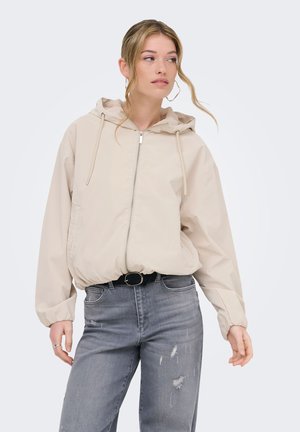 Jeune femme portant une veste à capuche beige zippée et un jean gris déchiré, regardant légèrement vers sa gauche avec des mèches de cheveux lâches.