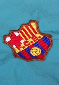 Geborduurde FC Barcelona-embleem met rode en gele strepen, een blauw en rood geblokt patroon, en een voetbal, op een teal-kleurige stof achtergrond.