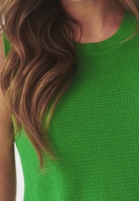 Lunghi capelli castani mossi sopra una maglia senza maniche verde brillante a maglia lavorata, che mostra il collo e le spalle.