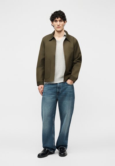 Jeune homme debout portant une veste vert olive, un pull gris clair, un jean bleu ample et des chaussures noires, sur un fond clair uni.