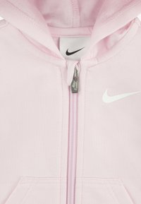 Rosa hettegenser med glidelås laget av mykt materiale. Har glidelås foran, to lommer og en hvit Nike-logo på brystet.