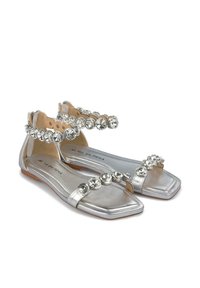 Silberne flache Sandalen mit einer viereckigen Zehenpartie, die entlang der Riemen mit klaren Schmucksteinen verziert sind und eine glatte metallische Textur aufweisen.