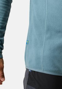Pullover in pile blu realizzato in materiale morbido, con superficie testurizzata, dettagli delle cuciture e una piccola bandierina gialla e blu sul lato.