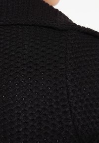 Svart stickad jacka med en strukturerad, våffel-liknande mönster. Har en bred krage och synliga sömmar längs sömmarna. Inga stängningar syns.