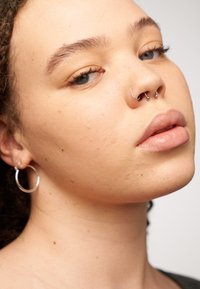 Gros plan d'un visage avec une peau brun clair. Présente plusieurs piercings faciaux en argent. Peau texturée, maquillage naturel et cheveux tressés.