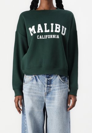 Osoba ubrana w ciemnozieloną bluzę z białym napisem „MALIBU CALIFORNIA” oraz jasnoniebieskie jeansy z wysokim stanem, stojąca na białym tle.