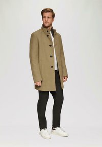 Khaki wollen jas met een staande kraag, zwarte knopen en zijzakken, gedragen over een grijze trui en zwarte broek, gecombineerd met witte schoenen.