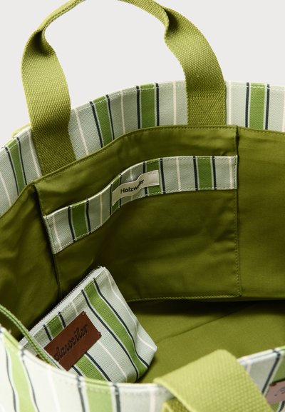 Εσωτερικό τσάντας με πράσινες και λευκές ρίγες, που δείχνει υφασμάτινες λαβές, εσωτερικές τσέπες και μια μικρή τσέπη με φερμουάρ με καφέ ετικέτα.