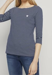 Navy und weiß gestreifte Langarmbluse mit einem Bootsausschnitt und einem kleinen weißen Herzlogo auf der linken Brust. Geeignet für Freizeitmode.