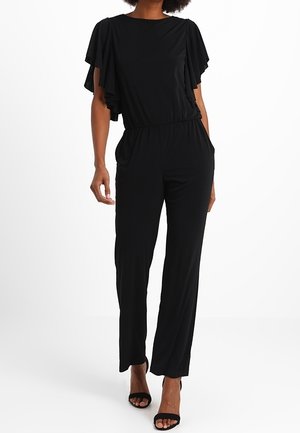 Vrouw draagt een zwarte jumpsuit met vliegermouwen, elastische taille, zijzakken en zwarte open-toe hoge hak sandalen.