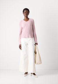 Lichtroze gebreide trui met een V-hals en uitlopende mouwen, gecombineerd met witte wijde broek en beige slingback hakken. Houdt een beige tas vast.
