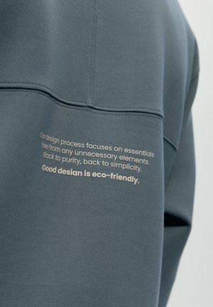 Nahaufnahme eines blauen Kleidungsstücks mit gedrucktem Text, der auf wesentliche, einfache und umweltfreundliche Designprinzipien auf dem Stoff nahe der Naht hinweist.