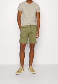 Beige t-shirt med et lille logo, olivengrønne shorts og lysebrune sneakers med hvide accenter. Enkel, afslappet outfit på en neutral baggrund.