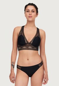 Naisterahvas mustas pitsist rinnahoidjas ja sobivates mustades bikiinialustes, seisev lihtsa heleda tausta ees.