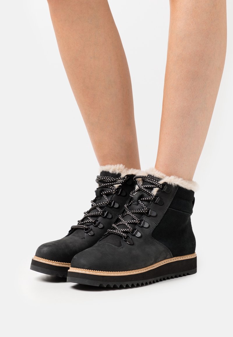 Bottes de randonnée en daim noir avec col en fourrure, lacets noirs et blancs, semelle en caoutchouc et crampons en zigzag pour une meilleure adhérence.