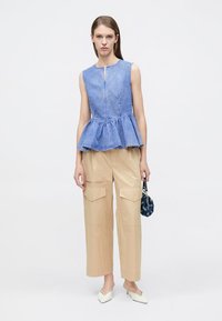 Μπλε ριγέ τοπ πέπλουμ με φερμουάρ στο μπροστινό μέρος, συνδυασμένο με μπεζ παντελόνι καμπάνα cargo με τσέπες. Το μοντέλο κρατάει μια τσάντα με σχέδιο.