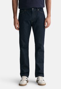 Jeans in denim scuro con una vestibilità dritta, dotati di tasche frontali, chiusura con bottone e cuciture a contrasto. Indossati con scarpe sportive di colore chiaro.