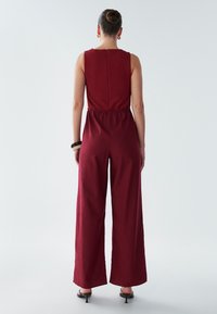 Combinaison bordeaux avec un haut smocké et un pantalon large. Fermeture éclair dans le dos, texture de tissu lisse, design minimal. Sans motifs ni accents.