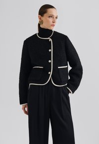 Veste noire texturée avec piping crème, col arrondi et fermetures à bouton. Comprend deux poches avant et est assortie à un pantalon noir.