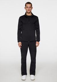 Svart zip-up jacka med långa ärmar, i kombination med svarta byxor som har sidofickor. Båda har en slät textur. Vita sneakers.