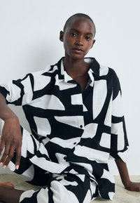 Majica s črno-belim vzorcem velikih abstraktnih oblik, izdelana iz lana, z ohlapnim krojem in zapenjanjem na gumbe, v kombinaciji s usklajenimi hlačami.