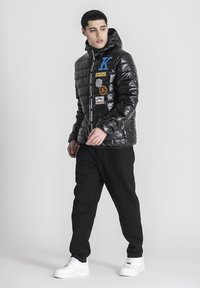Chaqueta acolchada negra con capucha, que presenta letras azules y varios parches. Llevada con pantalones negros y zapatillas blancas.