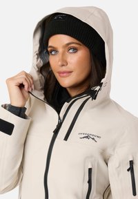 Giacca impermeabile beige con cappuccio, dotata di inserti neri con zip e logo. Il modello indossa un berretto lavorato a maglia nero con una toppa.