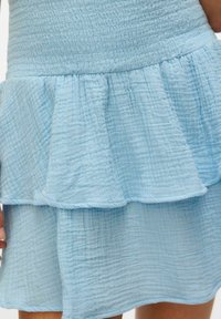 Abito azzurro chiaro con un tessuto testurizzato, caratterizzato da un orlo a balze a strati e un corpetto arricciato, che conferisce un aspetto morbido e fluido.