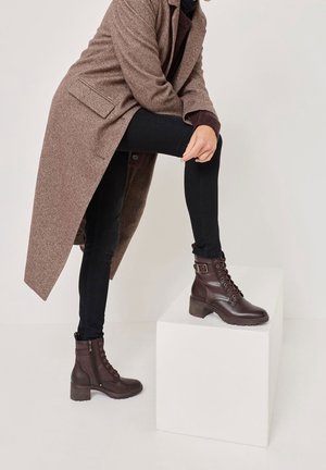 Hnědé kožené kotníkové boty se šněrováním vpředu, s přezkou a robustními podpatky. Nošené s tmavými džínami a dlouhým hnědým kabátem.