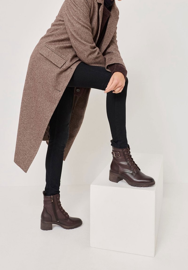 Bottes chevilles en cuir marron avec devant à lacets, détail boucle, et talons épais. Portées avec un jean foncé et un long manteau marron.