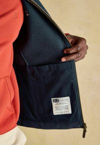 Jūras simbolikas jakas ar siltu flīsa oderi un zelta rāvējslēdzēju. Uz tās ir etiķete ar Lielbritānijas karogu un tekstu. Sarkanā piedurkne redzama fona.