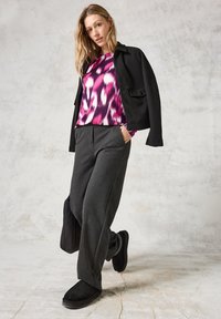 Schwarze Jacke über einem mehrfarbigen, gemusterten Hemd in Rosa- und Weißtönen, kombiniert mit grauen Hosen und schwarzen Slippern.