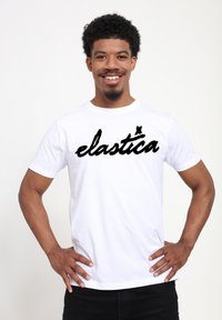 Bílá bavlněná tričko s černým kurzívním textem "elástica." Krátké rukávy, kulatý výstřih a volný střih.