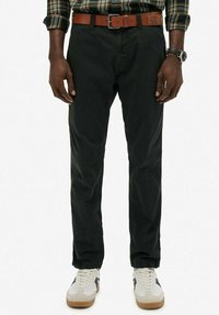 Mand iført sorte bukser, brunt læderbælte, beige og hvide sneakers, ur på venstre håndled og grøn ternet skjorte stoppet ind.