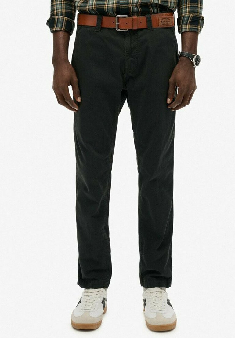 Un homme portant un pantalon noir, une ceinture marron, une chemise à carreaux avec les manches retroussées et des baskets blanches, se tenant contre un fond blanc.